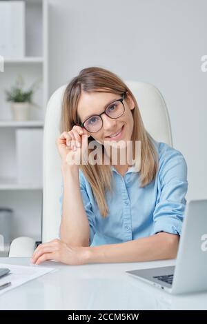 Happy young female office manager in eyeglasses and blue shirt looking at you Stock Photo
