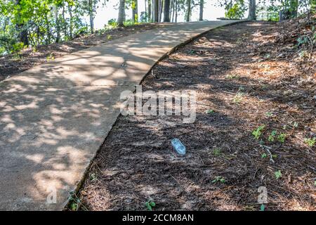 A plastic water bottle laying on the ground along the sidewalk with the garbage can being uphill on the trail in the woodlands of the park on a bright Stock Photo