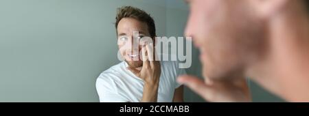 Man skin care putting moisturizer cream on face. Male beauty skincare concept. Young handsome adult looking in the bathroom mirror applying anti-aging Stock Photo
