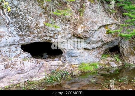 The entrance to a cave in the side of a cliff. A pool of water is in front of the opening. The cliff is made from limestone. Stock Photo