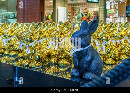 A black, glittering rabbit figure with a white ribbon stands in front of dozens of gold-coloured rabbit figures from the Easter bunny exhibition at the Stary Browar shopping mall in Poznań, Poland. Stock Photo