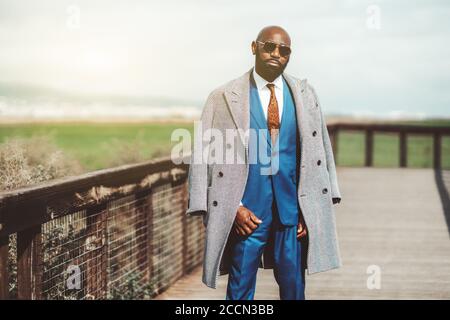 Portrait of a fashionable bald bearded mature black guy in an elegant blue costume with a necktie, striped coat, and sunglasses, he standing outdoors Stock Photo