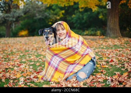 Woman wrapped in woollen blanket with dog in autumn fall park. Beautiful happy young Caucasian woman sitting on ground hugging domestic animal pet. Be Stock Photo