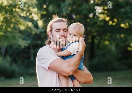 Proud father hugging embracing toddler baby boy outdoor. Young parent holding child son on arms. Authentic lifestyle tender moment. Single dad family Stock Photo