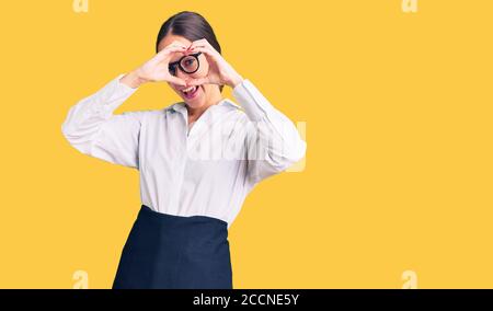 Beautiful brunette young woman wearing professional waitress apron doing heart shape with hand and fingers smiling looking through sign Stock Photo