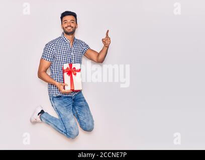 Young handsome hispanic man wearing casual clothes smiling happy. Jumping with smile on face holding birthday gift pointing with finger to the side ov Stock Photo