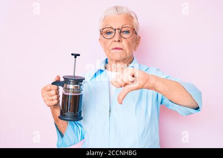 Senior beautiful woman with blue eyes and grey hair holding french coffee maker with angry face, negative sign showing dislike with thumbs down, rejec Stock Photo