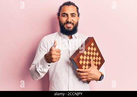 Young arab man holding chess smiling happy and positive, thumb up doing excellent and approval sign Stock Photo