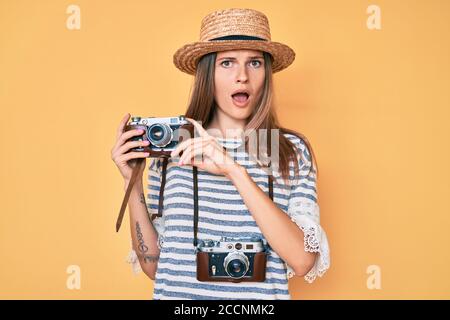 Beautiful caucasian tourist woman holding vintage camera in shock face, looking skeptical and sarcastic, surprised with open mouth Stock Photo