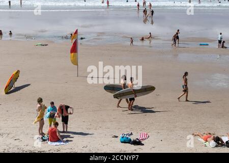 Beach day: two young men carrying surf table boards, family, kids playing, people walking, surf rescue table. Main beach, Byron Bay, Australia Stock Photo