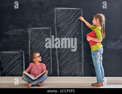 Back to school! Two happy cute industrious children indoors. Kid is drawing graphs on blackboard in the class. Girl reading a book. Stock Photo
