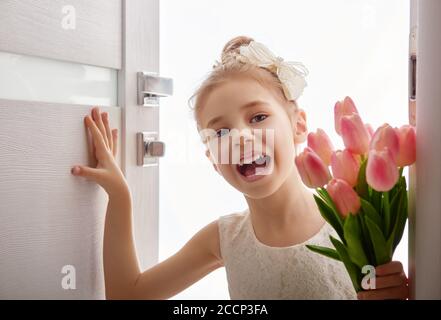Sweet child girl with bouquet of tulips. Happy little girl with bouquet of flowers opens the door. Wedding, Valentine concept. Stock Photo