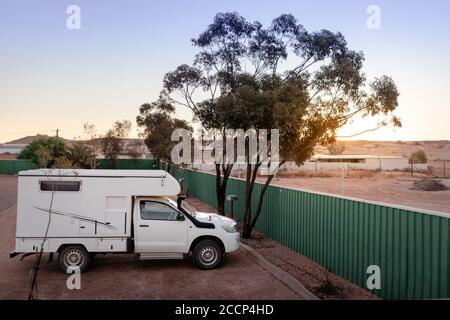 Small sized 4wd campervan parked on a camping site in the middle of the desert. Sunset time. Green fence and tree next to the car. White vehicle. Stock Photo