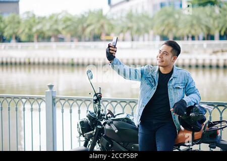 Happy handsome Vietnamese man standing at motorcycle, taking off helmet and taking selfie Stock Photo