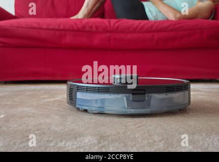 Robotic vacuum cleaner cleaning the room while woman relaxing on sofa Stock Photo
