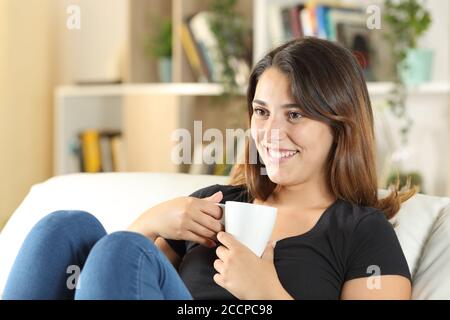 Happy woman holding coffee mug looks away sitting on a sofa in the living room at home Stock Photo