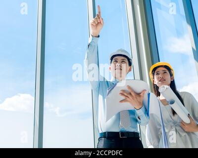 Asian architect Male and female inspecting construction work inside high-rise building near huge window glass on blue sky background. Couple engineer Stock Photo