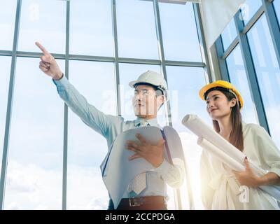 Asian architect Male and female inspecting construction work inside high-rise building near huge window glass on blue sky background. Couple engineer Stock Photo
