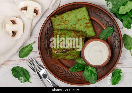 Spinach green pancakes (crepes) stuffed with mushroom filling with sour cream. Delicious healthy Breakfast in a clay plate on a light wooden backgroun Stock Photo