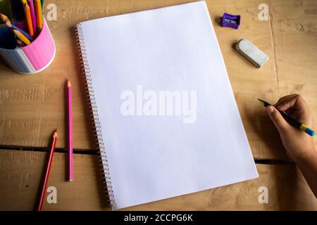 top view of an empty white notebook space for text, color pencils, eraser, sharpener and human hand holding pencil on wooden table Stock Photo