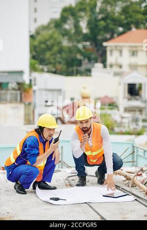 Head engineer checking construction plan when contractor controlling work of builders via walkie-talkie Stock Photo