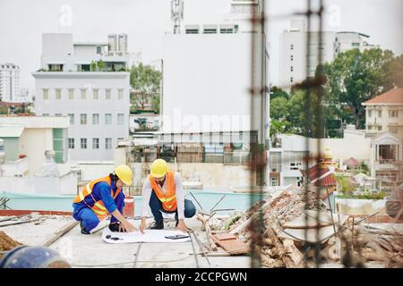 Builders in orange vests and hardhats pointing at building plan on the floor and discussing plan of works Stock Photo
