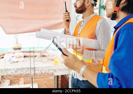 Civil engineer with blueprint talking on walkie-talkie when his colleague standing near by with digital tablet in hands Stock Photo