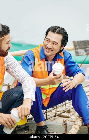 Handsome cheerful Vietnamese builder drinking coffee and talking to colleague during break Stock Photo