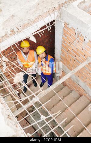 Builders with tablet computer stading on stairs in building under construction, talking on walkie-talkie and looking up Stock Photo