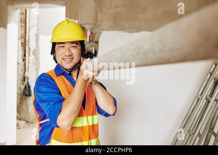 Portrait of smiling Vietnamese builder in bright orange vest and hardhat carrying metal pipe in building under construction Stock Photo