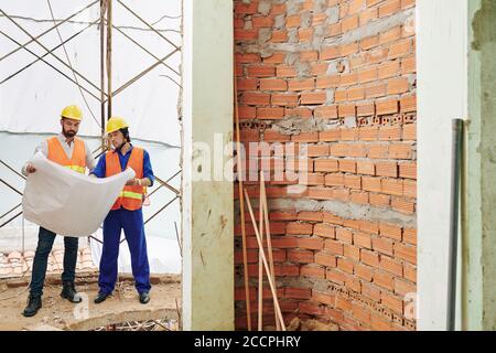Serious construction workers standing at unfinished brick wall and discussing construction plan Stock Photo