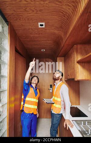 Electrician showing light sockets he installed in wooden ceiling in kitchen area to foreman with digital tablet in hands Stock Photo