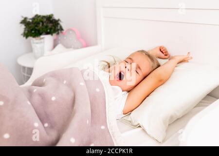 Top view of little cute blond girl sleeping on white bed with her hands up. Toddler yawns and don't want to wake up to go ro school. Stock Photo