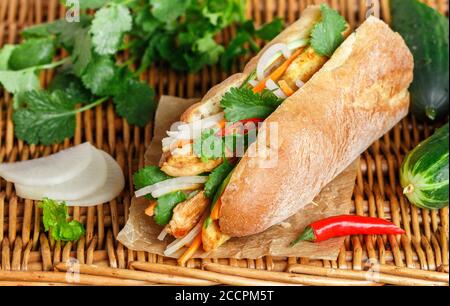 Baguette sandwich with fried chicken and fresh vegetables-carrots, cucumber, daikon, red pepper and cilantro (coriander) on a wicker table. Delicious Stock Photo