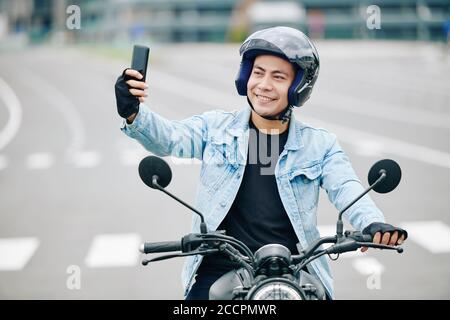 Happy handsome Vietnamese man in helmet taking selfie when sitting on his motorcycle Stock Photo