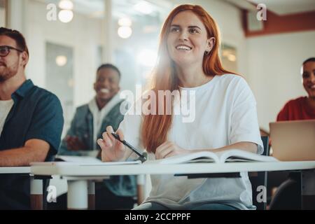 Pretty student paying attention in lecture. Girl listening to the teacher and smiling sitting in classroom. Stock Photo