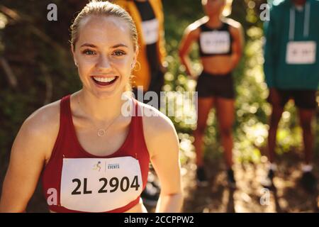 Woman runner standing outdoors looking at camera and smiling. Female in sportswear taking a break after a league run with athletes standing in backgro Stock Photo
