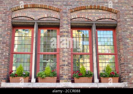 Two beautiful red windows on a brick facade with reflections on the windows glasses, there are potted flowers on the bottom Stock Photo