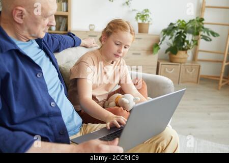 Portrait of cute red haired girl using laptop while enjoying time with grandfather in cozy home interior, copy space Stock Photo