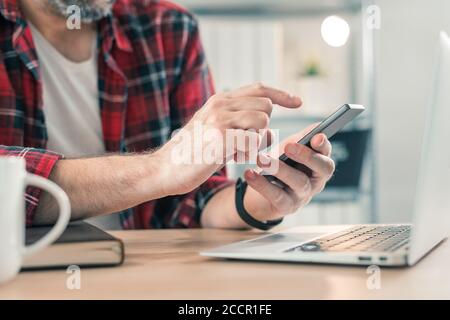 Freelancer using smartphone and laptop computer in home office while telecommuting, selective focus Stock Photo