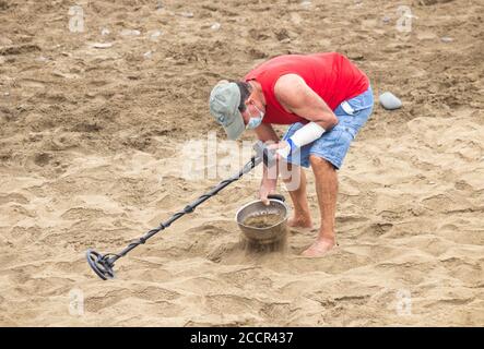 Man wearing face covering, face mask using metal detector on beach during Covid/Coronavirus pandemic. Stock Photo