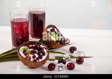 Dessert of cream and cherries and currants in coconut halves, two glasses of cherry juice and pieces of ice on a palm leaf on a white background. Copy Stock Photo