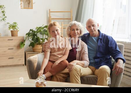 Warm toned portrait of happy senior couple posing with cute granddaughter while sitting on couch together in cozy home interior Stock Photo