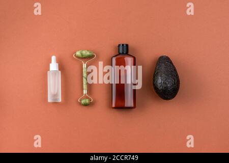 Brown bottle of lotion, serum or acid, jade face roller, avocado on brown surface. Cosmetic products and accessories creative composition. Natural Stock Photo