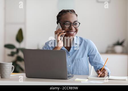 Black Female Office Manager Talking On Cellphone And Taking Notes At Workplace Stock Photo