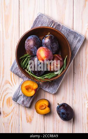 Fresh sweet plum fruits whole and sliced in brown wooden bowl with rosemary leaves on old cutting board, wood table background, top view Stock Photo