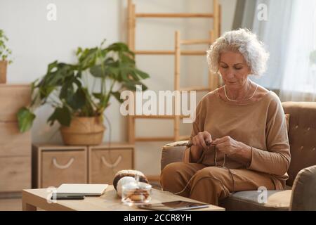 Warm toned portrait of content senior woman knitting in cozy home lit by sunlight, copy space Stock Photo