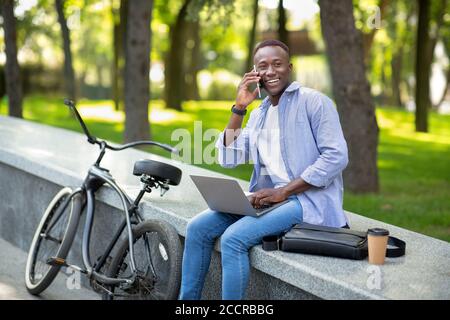 Happy black man with bicycle speaking on smartphone while using his laptop at park on summer day, copy space Stock Photo