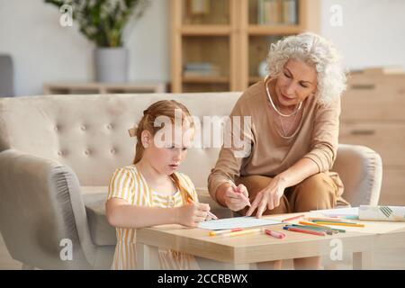 Warm toned portrait of senior woman babysitting cute red haired girl and drawing together while sitting in cozy living room Stock Photo