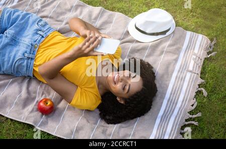 Cool black woman using mobile phone on picnic blanket outside, top view Stock Photo